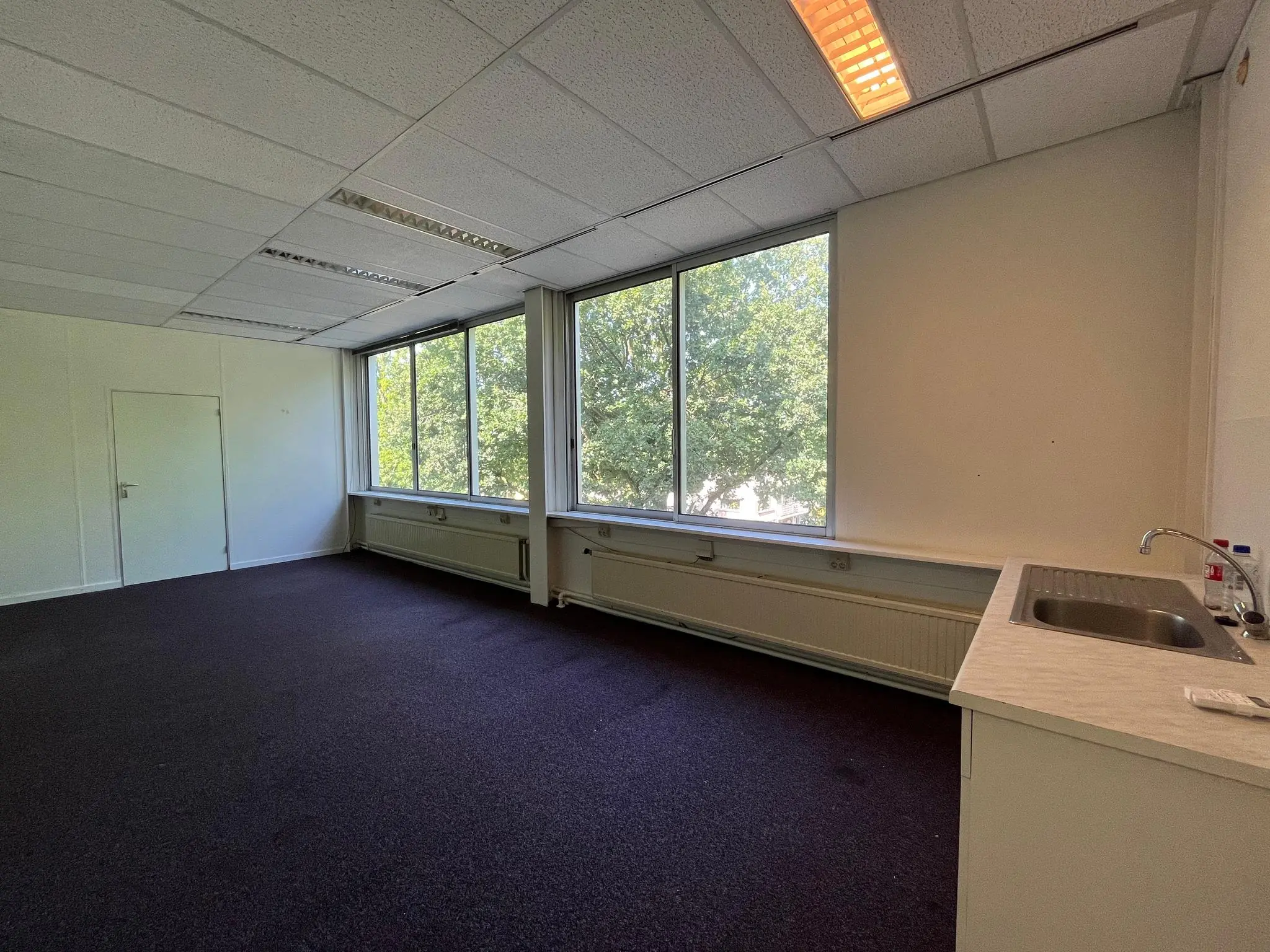 Leeg kantoorruimte aan de Jan van Eycklaan met grote ramen, een donker tapijt en een keukenblok met gootsteen.